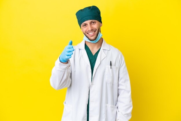 Chirurgo biondo in uniforme verde isolato su sfondo giallo che stringe la mano per chiudere un buon affare
