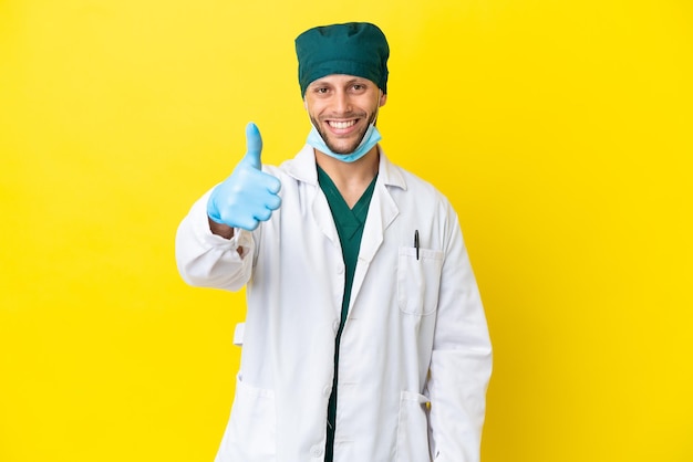 Chirurgo biondo in uniforme verde isolato su sfondo giallo con il pollice in alto perché è successo qualcosa di buono