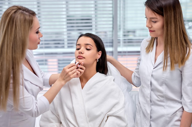 Chirurgo plastico caucasico professionista e assistente che disegna i contorni sul viso femminile con la matita prima...