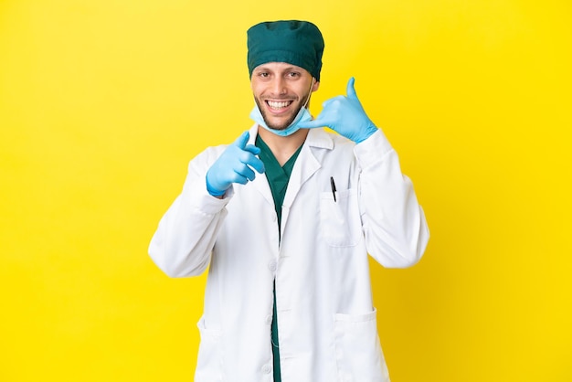 Chirurgo uomo biondo in uniforme verde isolato su sfondo giallo che fa il gesto del telefono e indica la parte anteriore