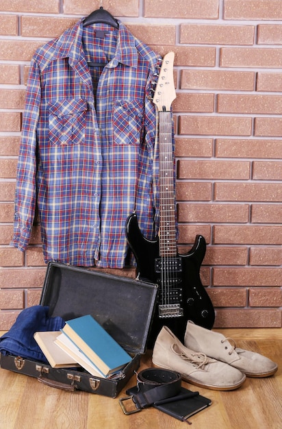 Chitarra vestiti stivali e valigia con libri sul pavimento sul fondo del muro di mattoni