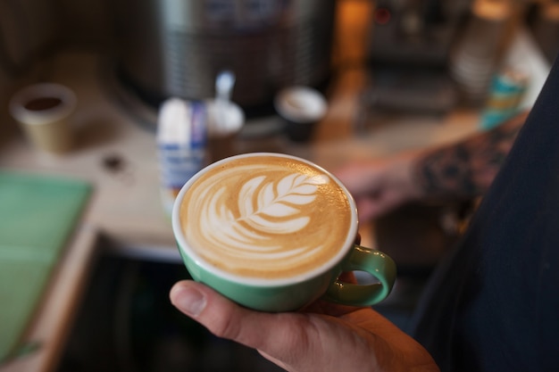 Chiuda in su del barista che tiene cappuccino aromatico. Caffè pronto per la vendita. Mani maschii che tengono una tazza di caffè.