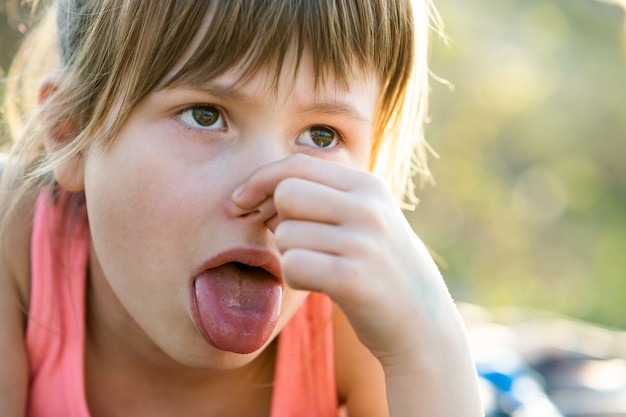 Chiuda in su del ritratto bellissimo bambino ragazza pizzicando il naso e mostrando la lingua.