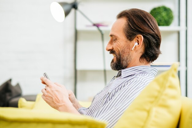 Chiuda in su dell'adulto caucasico allegro che si siede sul divano con gli auricolari wireless nelle orecchie e godendo utilizzando smartphone moderno