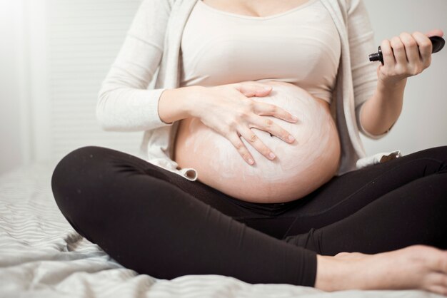 Chiuda in su della donna incinta che applica crema alla sua pancia