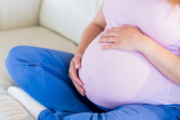 Chiuda in su della donna incinta che si siede sul divano che tocca la sua pancia