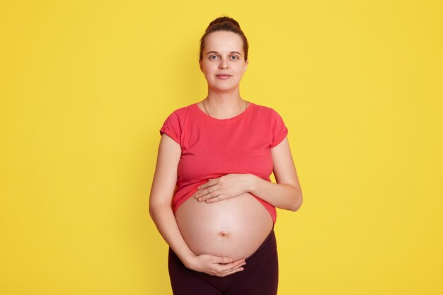 Chiuda in su della donna incinta che tocca la sua pancia nuda con entrambe le mani