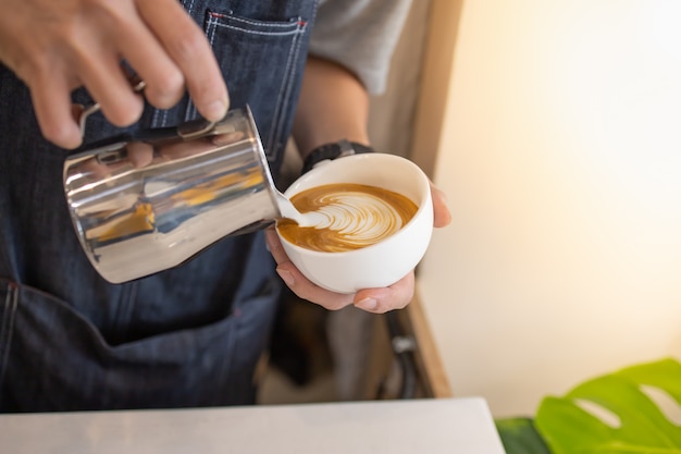 Chiuda in su della mano del barista che versa il latte stremato nella tazza bianca di caffè caldo per creare l&#39;arte del latte.