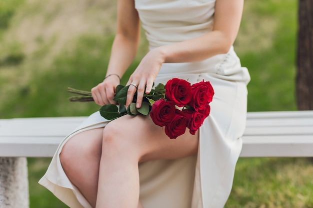 Chiuda in su della mano della giovane donna che tiene i fiori della rosa rossa su fondo all'aperto.