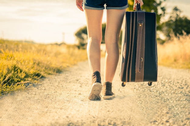 Chiuda in su delle gambe di una donna in vacanza che cammina sulla strada tenendo la sua valigia su uno sfondo di campagna