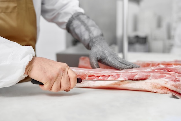 Chiuda in su delle mani del macellaio in guanti che tagliano la carne con la lama.