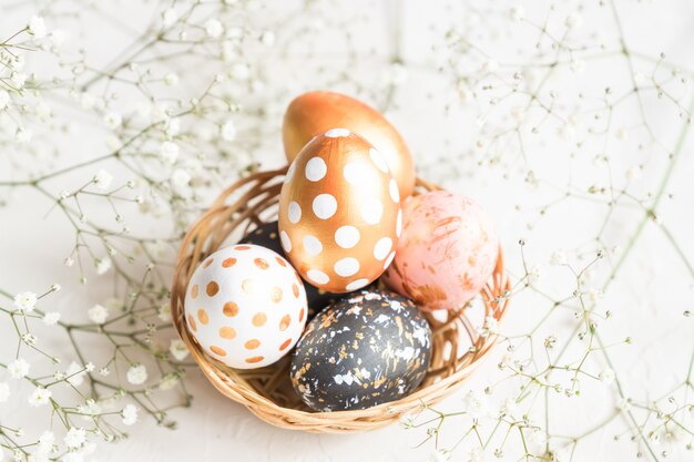 Chiuda in su delle uova di eeaster decorate nere, bianche e dorate in cestino di vimini decorato con il gypsophila su priorità bassa bianca.