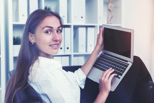 Chiuda in su di un allegro impiegato seduto al suo posto di lavoro con un computer portatile e digitando mentre guardando lo spettatore. Immagine tonica.