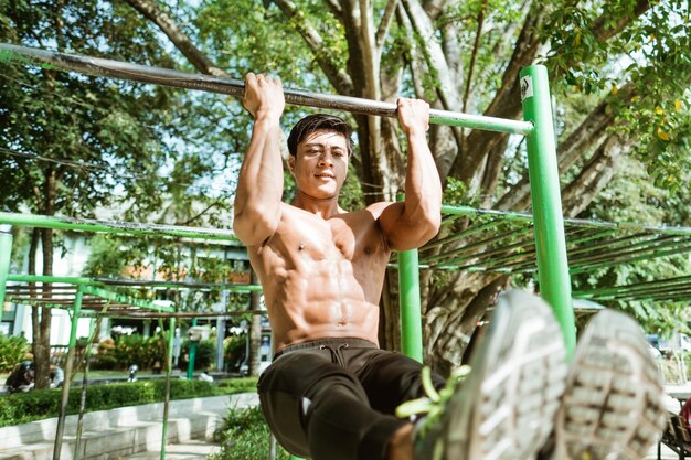 Chiuda in su di un uomo muscoloso asiatico senza vestiti facendo esercizi di pull-up L utilizzando la barra di ferro nel parco