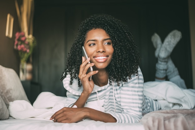 Chiuda in su di una donna di colore graziosa con capelli ricci che sorride e che per mezzo del telefono sul distogliere lo sguardo del letto