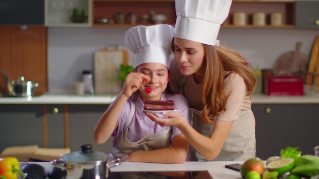 Chiuda in su piccolo pasticcere e madre che mette la ciliegina sulla torta in cucina moderna Madre che bacia la figlia a casa al rallentatore Due panettieri in uniformi del fornello che sorridono alla macchina fotografica sulla cucina