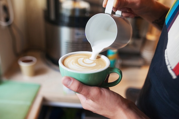 Chiuda in sulle mani del barista che versano latte caldo nella tazza di caffè per fare arte del latte. Arte del latte del caffè professionale nella caffetteria.