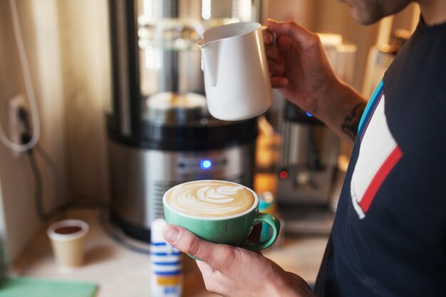 Chiuda in sulle mani del barista che versano latte caldo nella tazza di caffè per fare arte del latte. Arte del latte del caffè professionale nella caffetteria.