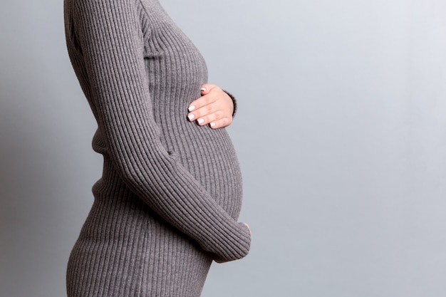 Chiuda su della donna incinta in vestito grigio che tiene la sua pancia al fondo grigio. Concetto di maternità. Copia spazio