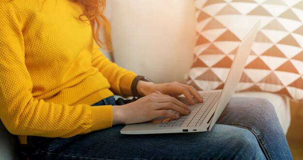 Chiuda su delle mani femminili con il computer portatile che si siede sullo strato in salone