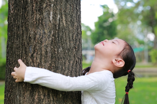 Chiuda su di un albero d&#39;abbraccio sorridente della ragazza del bambino con gli occhi chiusi al giardino.