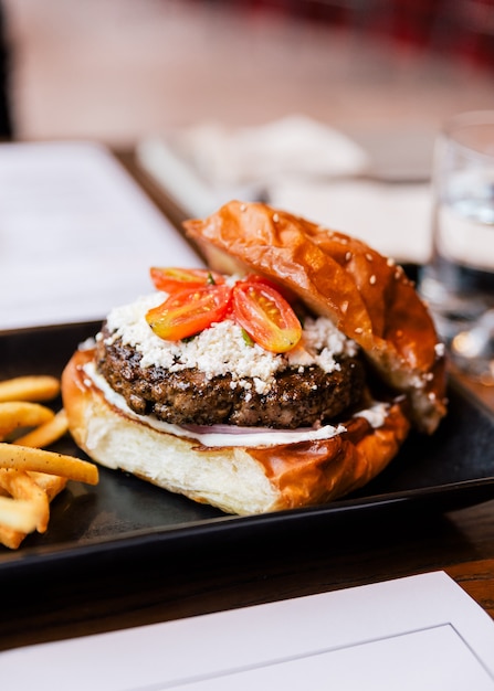 Chiuda sul cheeseburger con manzo grigliato, formaggio feta e pomodoro a fette servito con patatine fritte in lamiera nera.