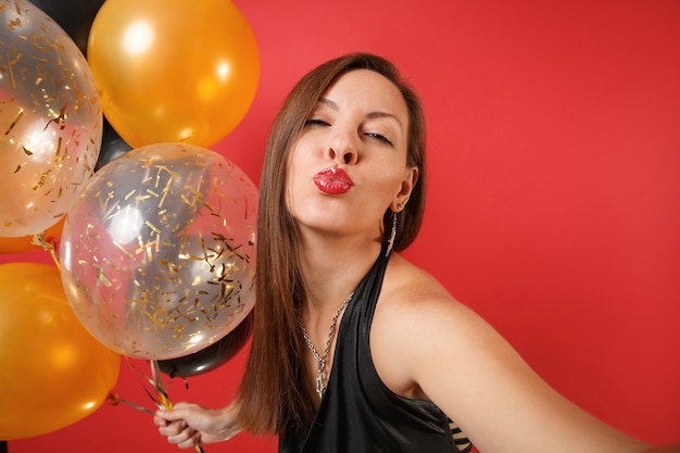 Chiuda sul colpo del selfie della donna in vestito nero che celebra il bacio di salto delle labbra tenere i palloni ad aria isolati su fondo rosso. Giornata internazionale della donna Felice Anno Nuovo compleanno mockup festa concetto.