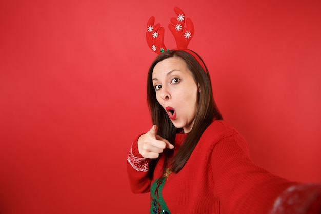 Chiuda sul colpo selfie della ragazza stupita della Santa in corna di cervo decorative che puntano il dito sulla macchina fotografica, tenendo la bocca spalancata isolata su sfondo rosso. Felice Anno Nuovo 2019 celebrazione festa concetto.