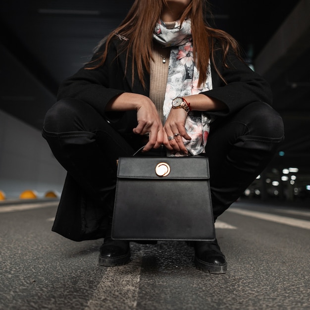 Chiuda sul corpo femminile in abiti stagionali neri eleganti con borsa in pelle vintage sulla strada in città. La ragazza attraente è seduta sull'asfalto in scarpe alla moda con borsa alla moda. Moda primaverile.