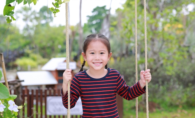 Chiuda sul piccolo gioco asiatico felice della ragazza del bambino e sedersi sull&#39;oscillazione nel parco naturale.