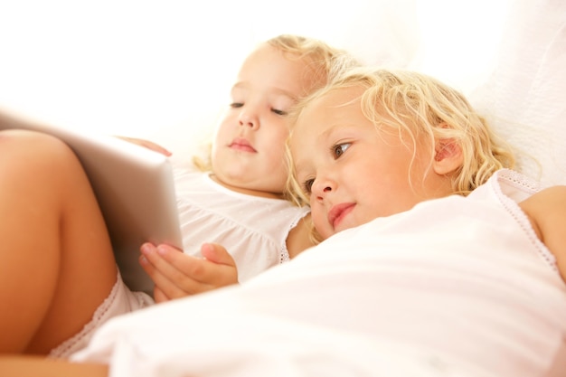 Chiuda sul ritratto dei bambini che utilizzano la compressa digitale sul letto