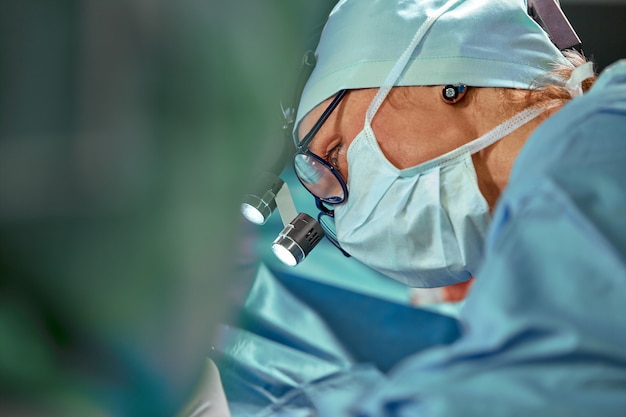 Chiuda sul ritratto del medico chirurgo femminile che indossa la maschera protettiva e cappello durante l'operazione.