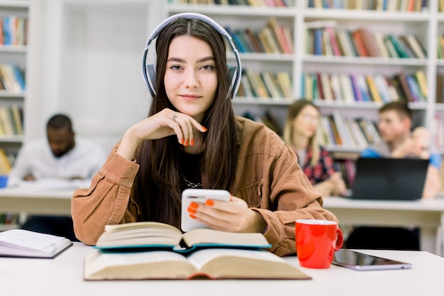 Chiuda sul ritratto dell'affascinante studente di ragazza sorridente carina, seduto in biblioteca al tavolo con molti libri, ascolto musica in cuffia