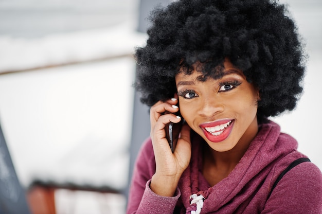 Chiuda sul ritratto della donna afroamericana dei capelli ricci posata al giorno di inverno, parlando sul telefono cellulare.