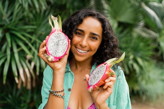 Chiuda sul ritratto della donna castana adorabile sorridente con i capelli ondulati che tengono i frutti tropicali