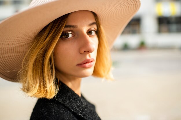 Chiuda sul ritratto della ragazza bionda alla moda elegante dei capelli corti in cappello che posa sopra il fondo della via