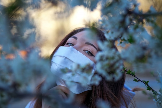 Chiuda sul ritratto della ragazza sotto un albero sbocciante con una maschera dal coronavirus.