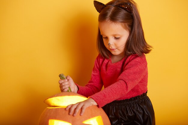 Chiuda sul ritratto della ragazza sveglia che gioca con la sua zucca di Halloween, il maglione da portare del bambino e la gonna nera, piccolo modello affascinante con le orecchie di gatto