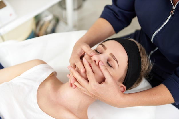 Chiuda sul ritratto delle mani dell'estetista che fanno un massaggio facciale ringiovanente professionale alla giovane donna bionda che si rilassa sul lettino da massaggio al salone della stazione termale. Concetto di cura della pelle