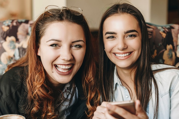 Chiuda sul ritratto di due donne adorabili che guarda l'obbiettivo ridendo mentre si tiene uno smartphone seduto in un caffè.