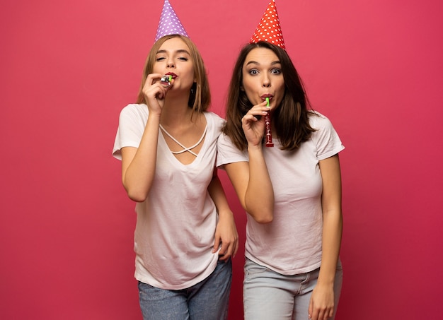 Chiuda sul ritratto di giovani donne bionde e castane con cappelli di compleanno che hanno divertimento isolato su sfondo rosa.