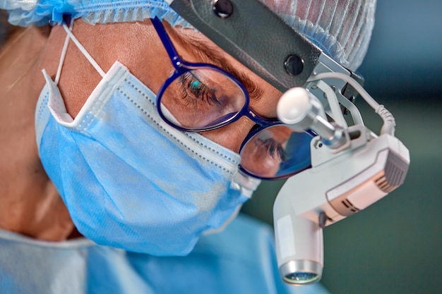 Chiuda sul ritratto di medico chirurgo femminile che indossa la maschera protettiva e cappello durante l'operazione. Assistenza sanitaria, educazione medica, concetto di chirurgia.