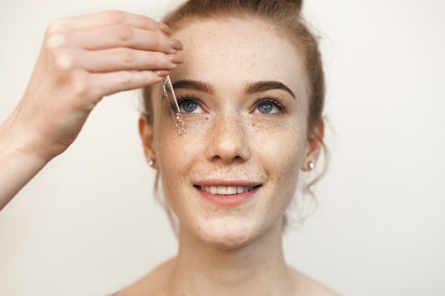 Chiuda sul ritratto di una bella donna dai capelli rossi che applica un siero trasparente con acido ialuronico sul viso sorridente isolato su bianco.