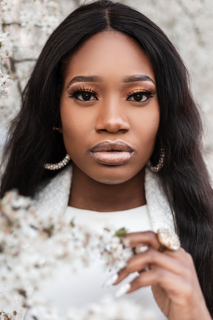 Chiuda sul ritratto di una bella donna di colore con vestiti bianchi in fiori all'aperto. Ragazza africana in natura in un giorno di primavera