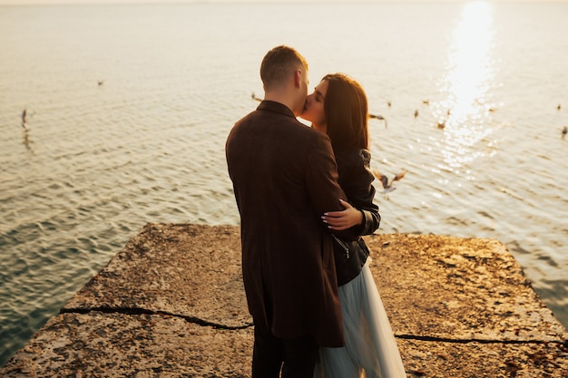 Chiuda sul ritratto di una bella giovane coppia elegante innamorata in piedi e si baciano sulla spiaggia al tramonto.