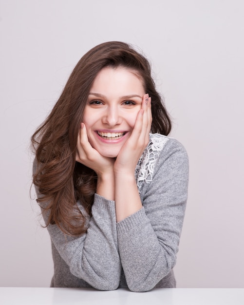 Chiuda sul ritratto di una donna sorridente felice che riposa il suo mento sulle sue mani e che esamina direttamente la macchina fotografica