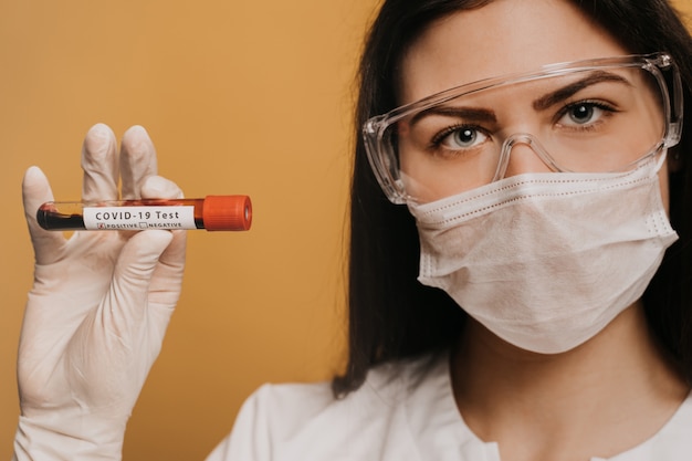Chiuda sul ritratto di una dottoressa in maschera chirurgica, guanti e occhiali protettivi in possesso di una provetta con un test Covid-19 coronavirus. Concetto di medici, infezione, ricerca e covid19.
