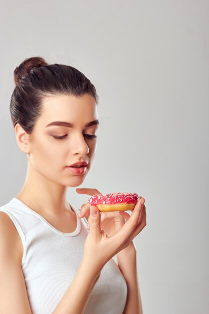 Chiuda sul ritratto di una ragazza affamata che mangia i dessert isolati. La ragazza castana tiene e gusta un dessert rosa