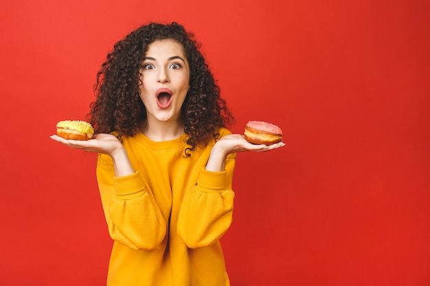 Chiuda sul ritratto di una ragazza graziosa soddisfatta che mangia le ciambelle isolate sopra fondo rosso.