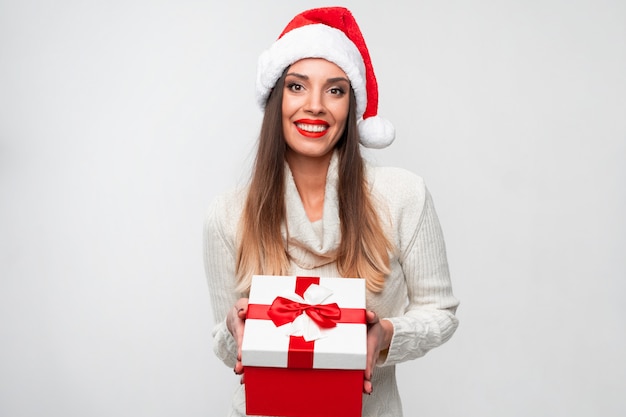 Chiuda sulla donna caucasica del beautifiul del ritratto in cappello rosso di Santa sulla parete bianca. Concetto di Natale Capodanno Sorpreso carino denti donna sorridenti emozioni positive dà grande confezione regalo rosso